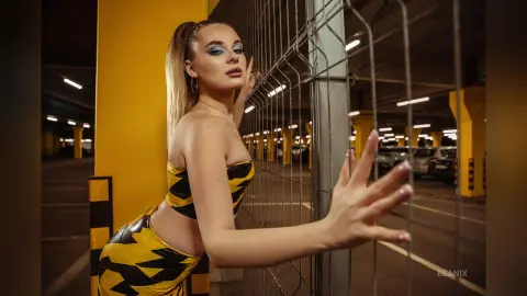 BeaNix poses against a metal wire fence in a yellow and black patterned bikini, looking over her shoulder with blonde hair in a high ponytail.