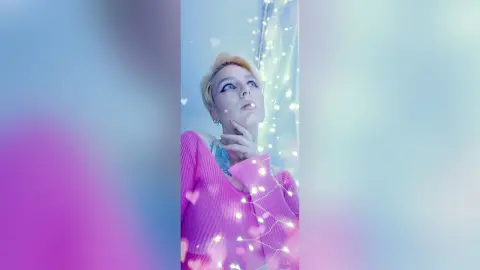 AvaLindt looks up with a hand to her chin, wearing a pink ribbed sweater against a soft, glowing light background.