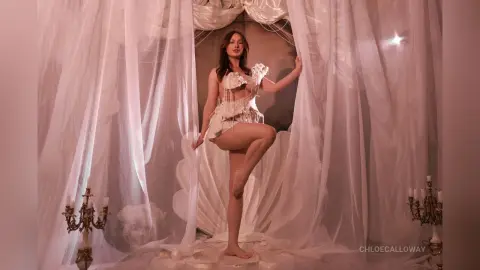 Elegant woman in a white lace bodysuit posing inside a dreamy, ethereal seashell set with flowing sheer curtains.