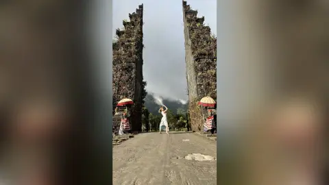 DaisyLin stands between two tall stone pillars wearing a white dress against a cloudy sky.