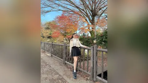 Smiling young woman in a white blouse and black skirt standing by a wooden fence under autumn trees.