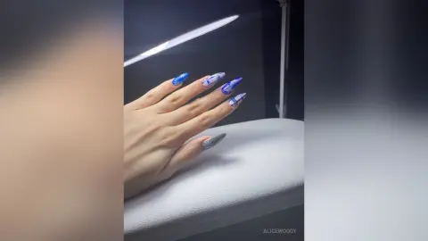 Close-up of long blue almond-shaped nails with white floral nail art designs on a white surface.