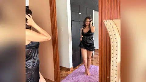 Young woman in a black satin slip dress standing in a bedroom looking into a full-length mirror.