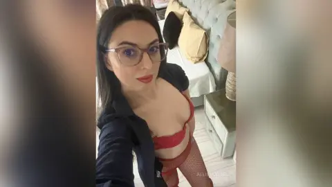 Young woman with dark hair and glasses wearing a black shirt and red lace lingerie in a bright bedroom.
