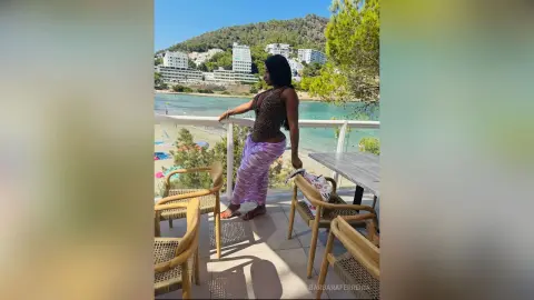 Stylish woman in a purple skirt and patterned top relaxing on a sunny Mediterranean seaside balcony.