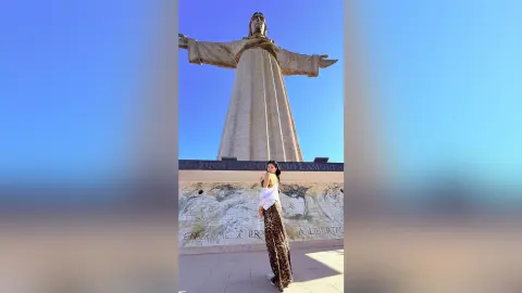 Woman in a white top and leopard print skirt standing before a massive Christ statue under a clear blue sky.