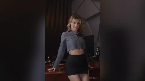 Smiling woman in a grey button-down shirt and black mini skirt posing in a modern office setting.