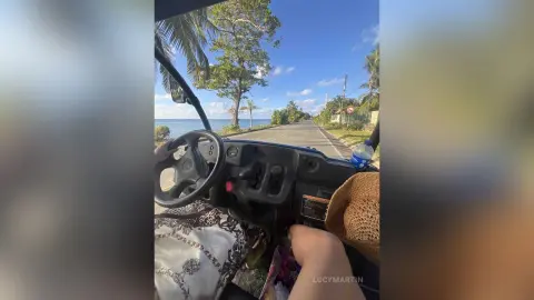 LucyMartin sits behind the steering wheel of a golf cart on a paved road, wearing a white patterned top with a view of her bare legs.