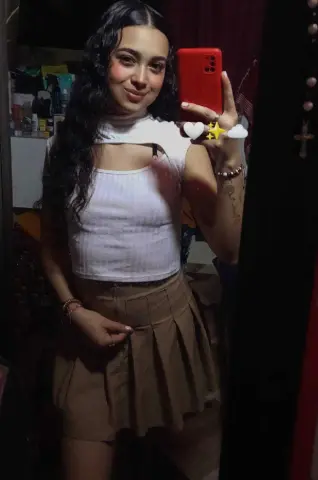 Young woman with wavy hair in a white ribbed crop top and tan pleated skirt taking a mirror selfie.