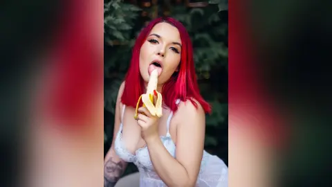 Young woman with vibrant red hair in a white lace outfit eating a banana in a lush green garden setting.