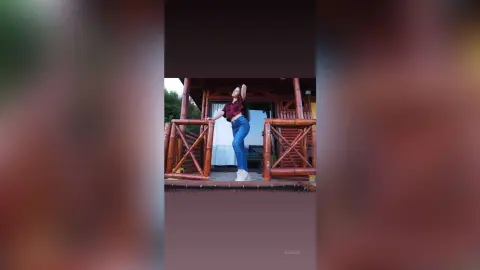 JessieLiu stands on a wooden deck wearing a maroon top and blue jeans with one arm raised.