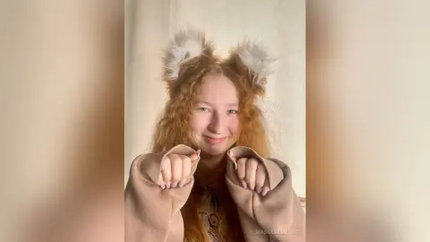 Smiling redhead girl wearing fluffy animal ears and a cozy beige sweater in a bright indoor setting.