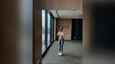 Fit woman in grey leggings and white crop top taking a mirror selfie in a modern minimalist gym setting.