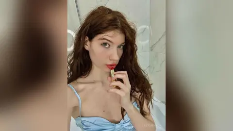 Young woman with wavy brown hair applying red lipstick in a bright marble bathroom wearing a light blue camisole.