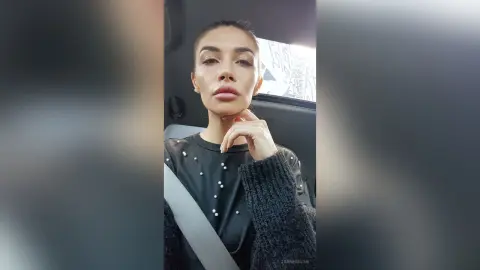 JanneMaine looks directly at the camera wearing a black long-sleeved top with white pearl-like studs, sitting in a car.
