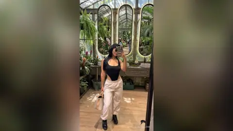 Woman in black bodysuit and beige trousers taking a mirror selfie in a lush botanical garden greenhouse.