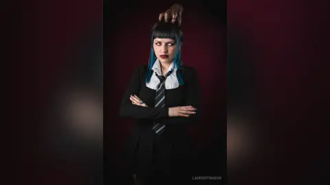 Moody portrait of a woman with blue hair wearing a black dress and striped tie against a dark background.