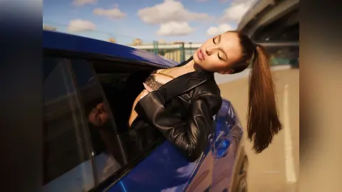 DelanaGrace leans out of a blue car window wearing a black leather jacket and black lace bra, her long hair in a high ponytail.
