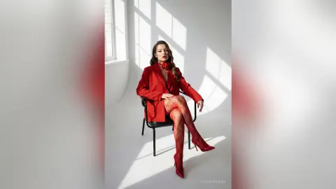 Elegant woman in a red blazer and patterned tights sitting in a bright, minimalist studio setting.