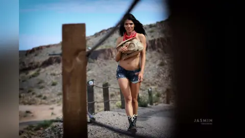 DrellaWolf — Beautiful brunette woman in denim shorts and cowboy boots holding a straw hat in a sunny desert landscape.