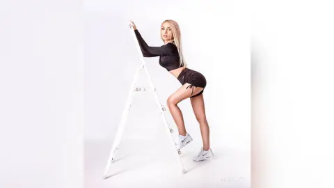 MonaLys poses in a black long-sleeve crop top and black shorts while leaning against a white ladder against a white background.