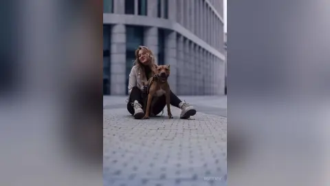 InaStark sits on a city sidewalk with her brown dog, wearing a white jacket and black leggings.