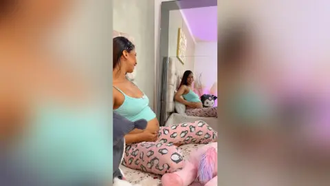 Smiling pregnant woman in a mint tank top and leopard print pajama pants relaxing in a cozy bedroom.