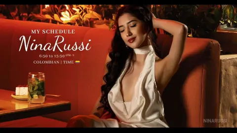 Nina Russi in a white satin halter dress sitting on a warm orange sofa in a moody, dimly lit setting.