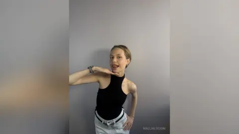MalliaLucioni poses in a black halter top and white pants, sticking her tongue out against a plain grey wall.