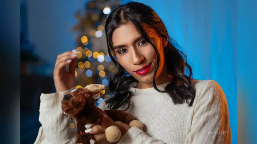 AnnMyer — Smiling young woman in a cozy white sweater holding a brown teddy bear in a warm, festive setting.