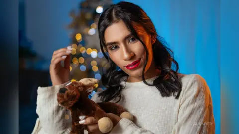 Smiling young woman in a cozy white sweater holding a brown teddy bear in a warm, festive setting.
