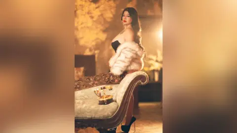 Elegant woman in a white faux fur coat sitting on a vintage chaise lounge in a warm, moody indoor setting.