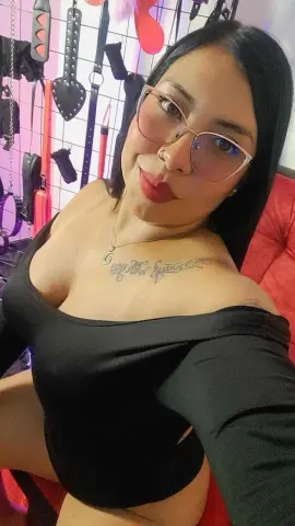 Smiling woman with glasses and tattoos wearing a black off-the-shoulder top in a boutique setting.