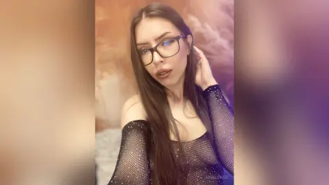 Young woman with long brown hair and glasses wearing a black mesh top in a soft, moody indoor setting.
