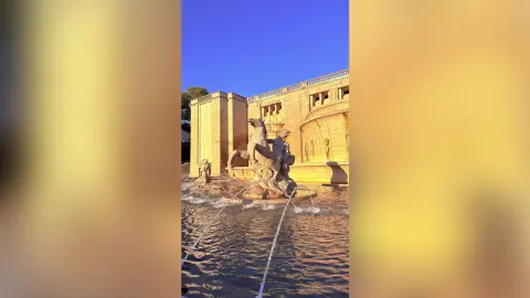 Classical stone fountain sculpture with water flowing in front of a grand historic building at golden hour.