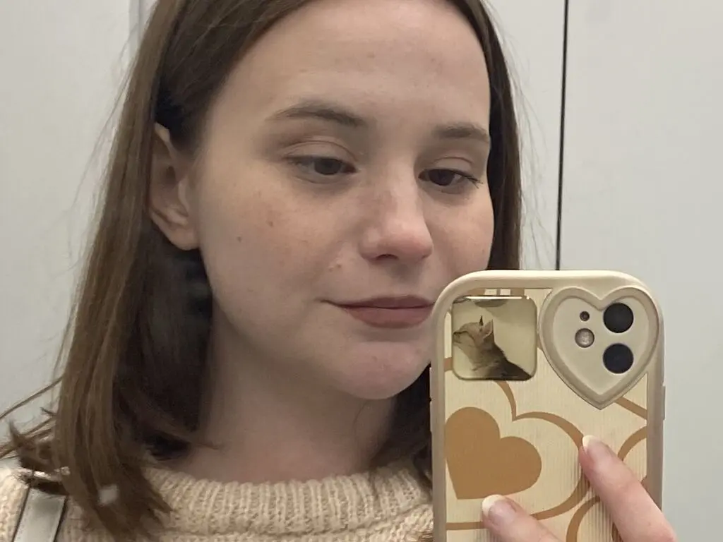Young woman with long brown hair wearing a beige knit sweater taking a casual selfie in a bright room.