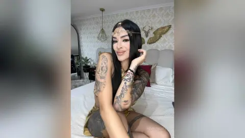 Tattooed woman in gold goddess costume and headpiece sitting on a bed in a bright, elegant bedroom.