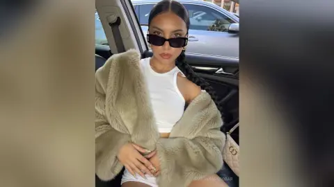 Stylish woman in a white tank top and beige faux fur coat wearing sunglasses sitting in a car.
