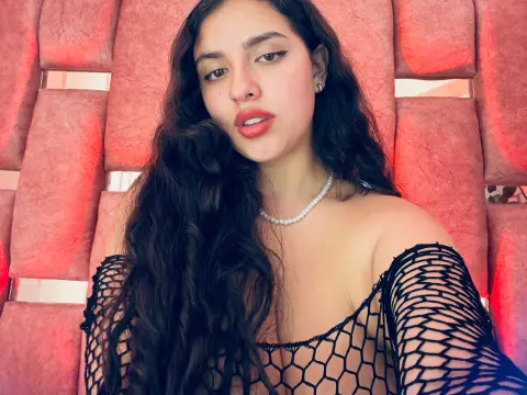 Young woman with long wavy hair wearing a pearl necklace and black mesh top against a pink textured background.