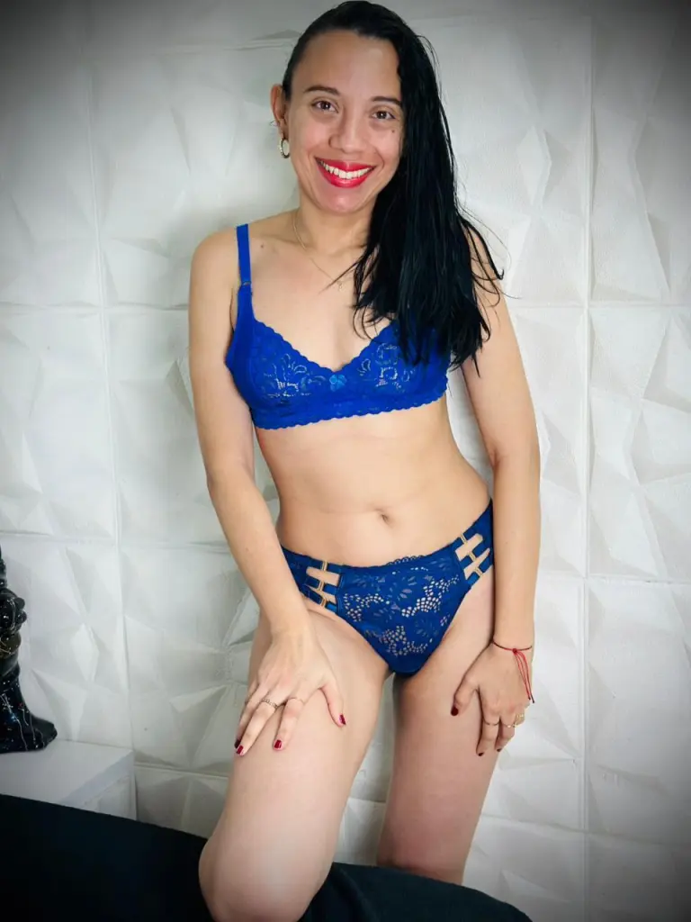 PerlaJohnsonRx with long dark hair, wearing a grey crop top, posing in a bright room.