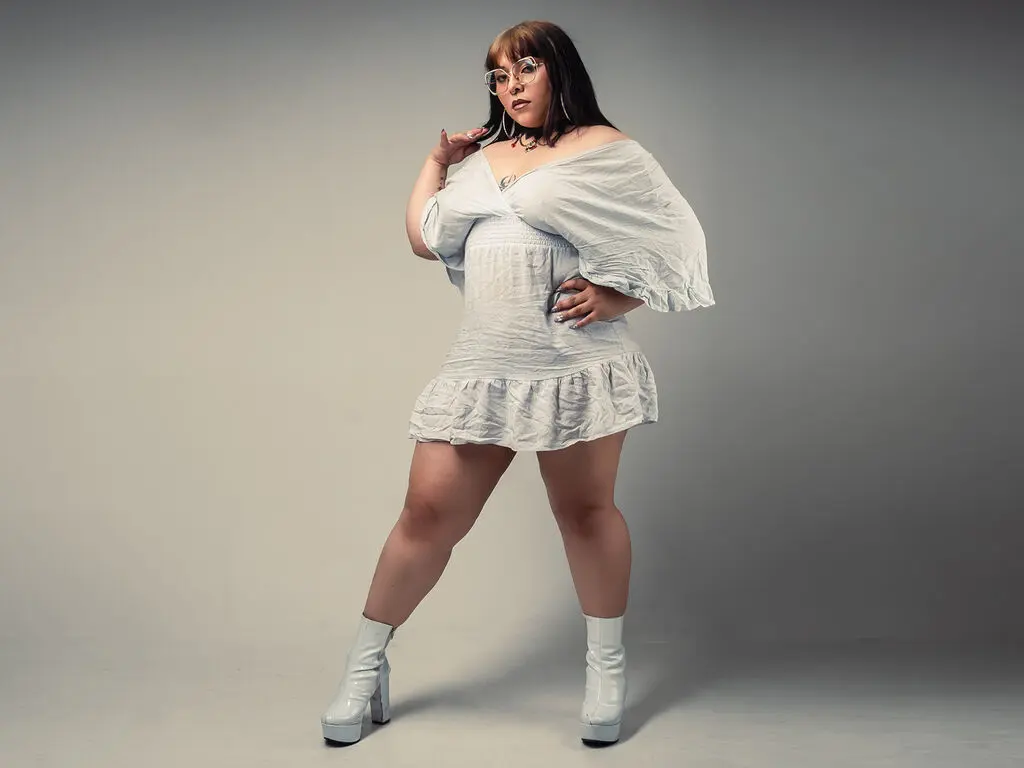 Stylish woman in a white mini dress and platform boots posing confidently against a grey studio background.