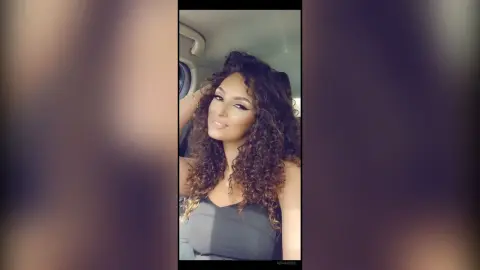 Smiling woman with voluminous curly hair wearing a grey top inside a car during golden hour.