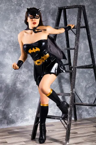 Woman in black Catwoman cosplay costume posing with a ladder against a textured grey studio background.