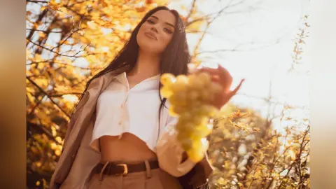 Beautiful woman in a white crop top holding green grapes in a sunlit autumn vineyard.