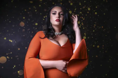 Elegant woman in an orange evening gown and diamond necklace posing against a dark bokeh background.
