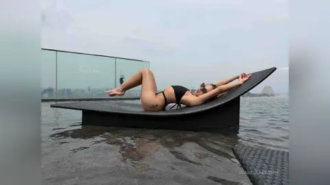 Relaxed woman in black bikini lounging on a sunbed by a rooftop infinity pool under a cloudy sky.