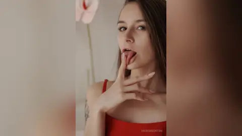 IngridBeckton licks her finger and touches her lips while looking at the camera, wearing a red tank top with her long brown hair down.