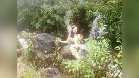 Smiling woman in a floral bikini relaxing by a lush jungle waterfall amidst tropical greenery.