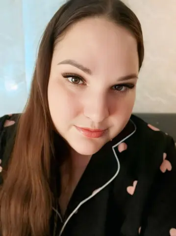 Close-up portrait of a smiling woman with long brown hair wearing black heart-patterned pajamas in a bright room.