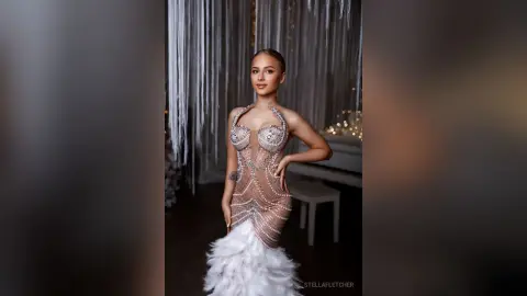 StellaFletcher stands wearing a sheer, crystal-embellished bodysuit with a feathered skirt against a silver tinsel backdrop.
